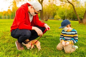 Young caucasian father and son in medical protective masks stay in park at sunny day on grass. Dad and child boy walk and play with the cute little puppy dog chihuahua