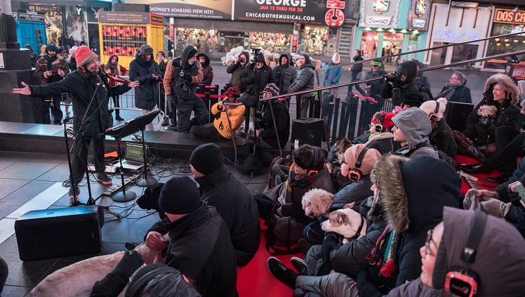 Laurie Anderson stands on stage with her arms open. An audience of humans and dogs huddle together to stay warm.