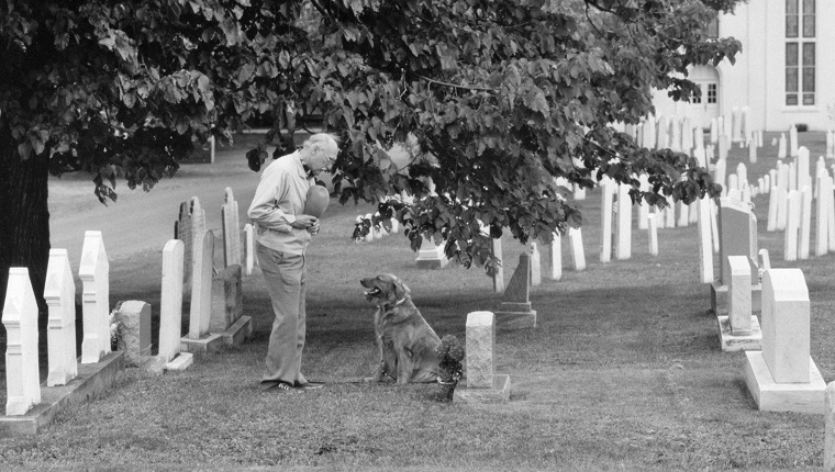 ELDERLY MAN HAT OVER HIS HEART AND PET DOG VISITING GRAVE OF LOVED ONE IN CHURCHYARD CEMETERY (Photo by Camerique/ClassicStock/Getty Images)