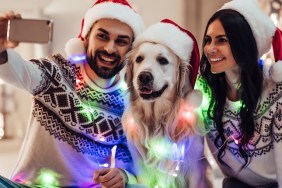 Merry Christmas and Happy New Year! Happy couple with dog labrador retriever waiting for the New Year in Santa Claus hats while sitting near beautiful Christmas tree at home. Smiling and making selfie