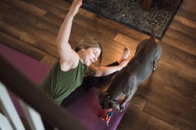 An adult woman does yoga and strength training exercises on a mat in her living room, her pet dog keeping her company and trying to play.