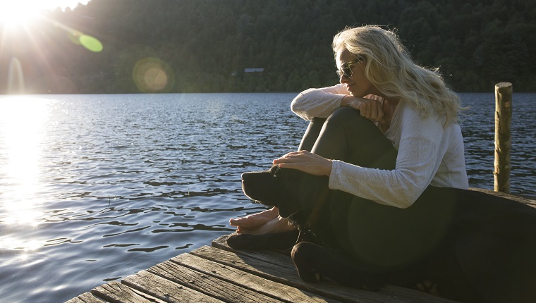 Woman pats dog on lake pier, sunrise