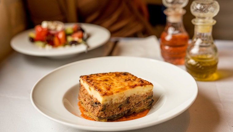 Slice of Greek moussaka served on a plate with Greek salad as a side dish white tablecloth with olive oil and vinegar in a restaurant