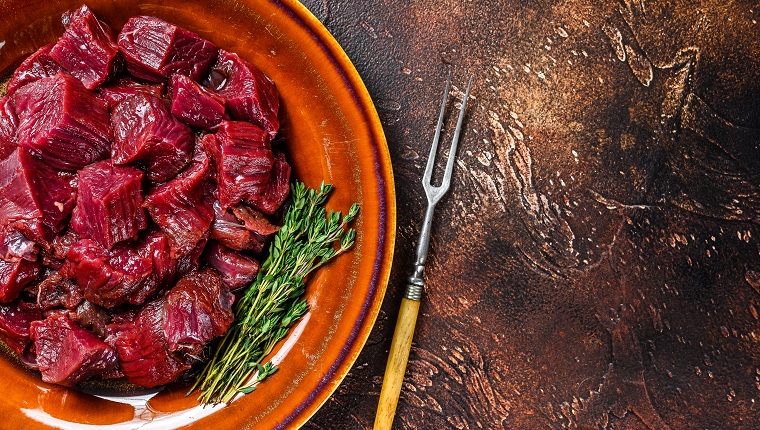 Raw diced elk meat steak for a goulash on a plate. Dark background. Top view. Copy space.