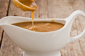 A close up shot of a small white ladle pouring brown gravy into a white ceramic gravy boat. Shot on a grungy old wooden table.