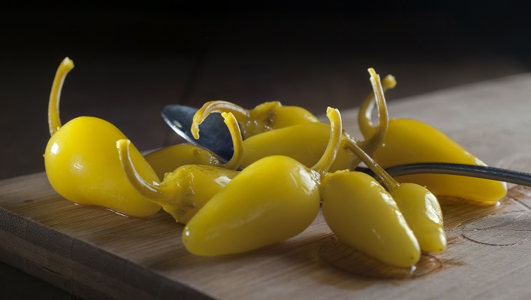 pepperoncini peppers on a spoon and on a wooden cutting board macro photography or close up image, in a dark background spicy food concept ready for salad preparation.