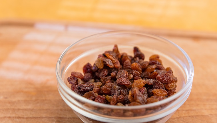 Bowl of raisins. Sultana raisins in glass bowl. Wooden chopping board. Yellowish background. Light effect. Selective focus.