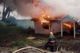 A woman firefighter works to move equipment, a burning unrecognizable home fully engulfed in fire and smoke in the background.